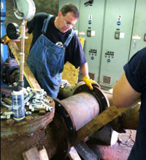 Man in overalls repairing industrial equipment, wearing gloves. Inside, near a control panel.