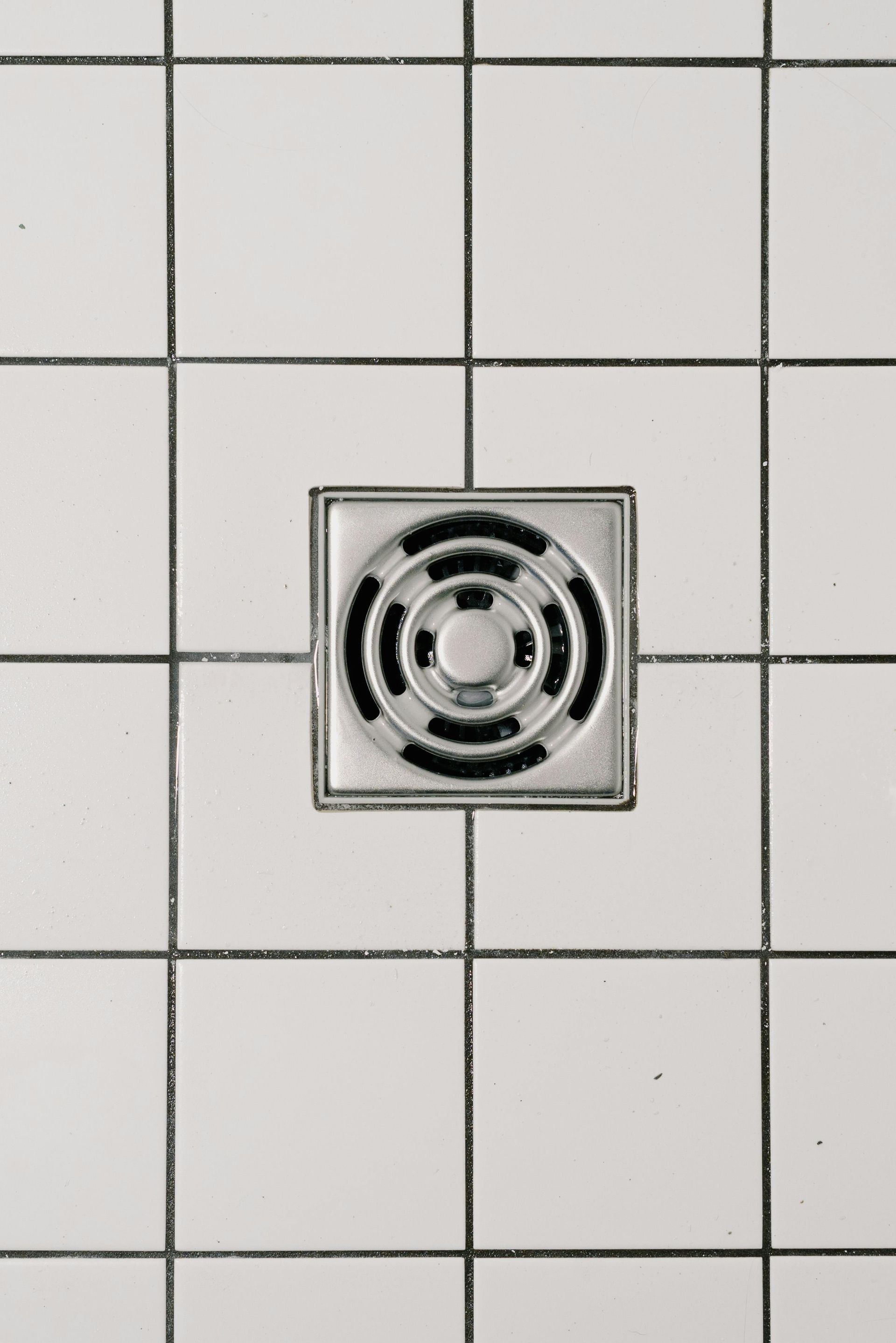 Square shower drain in white tiled floor with black grout.