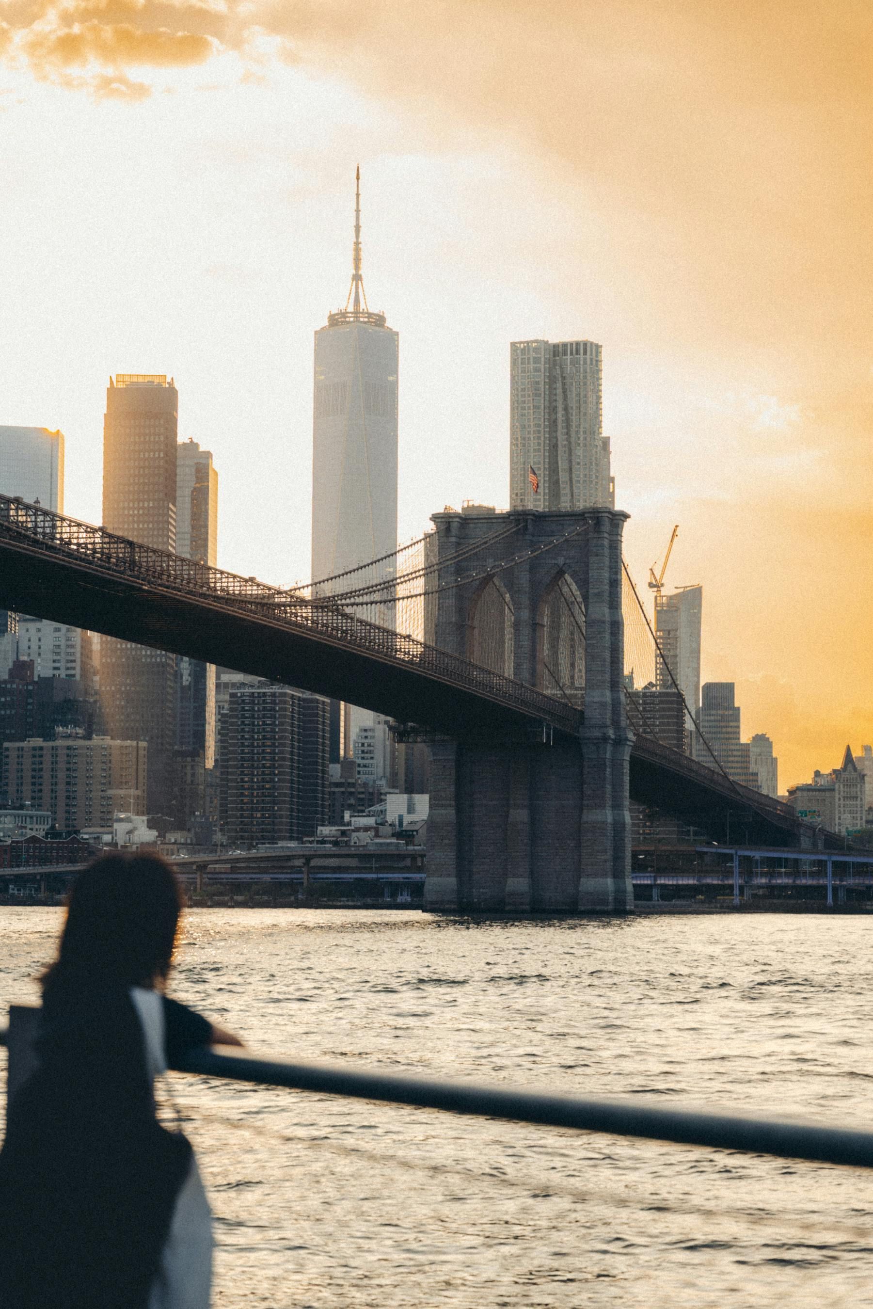 A person looks across the water at the Brooklyn Bridge and the New York City skyline during a golden sunset.