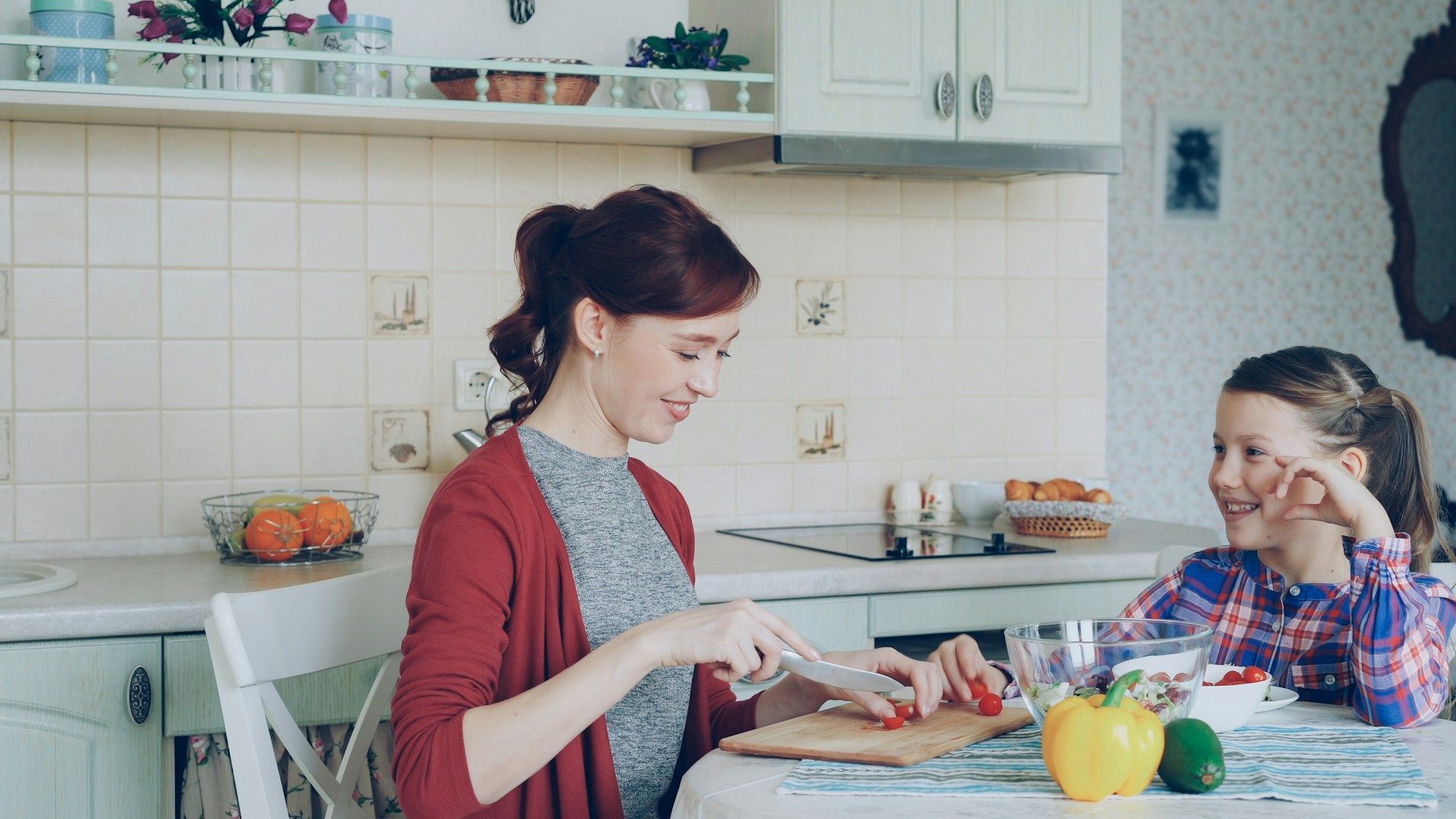 Woman in red cardigan slices tomatoes, child watches at a table in the kitchen.