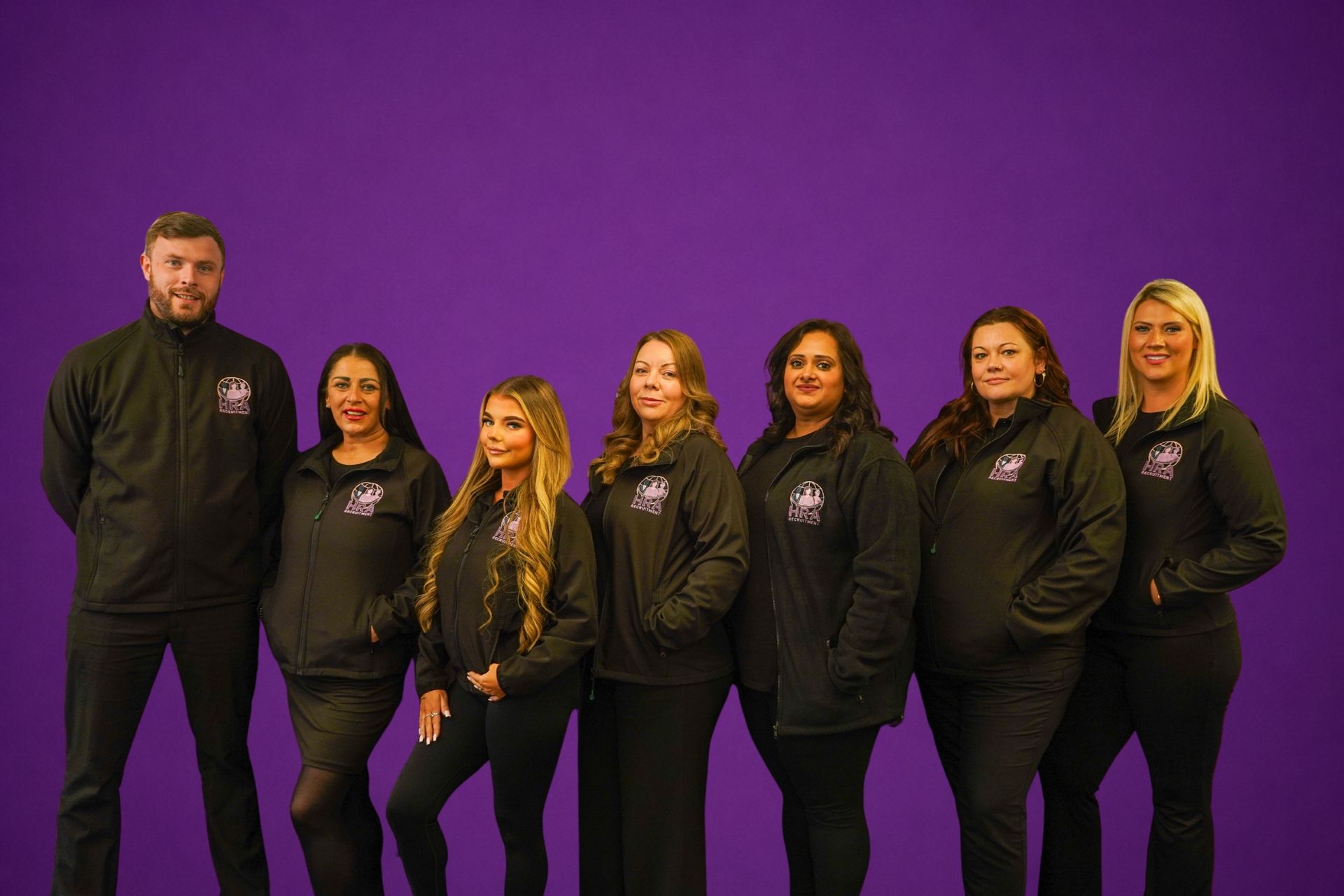 Seven people in black hoodies posing against a purple backdrop