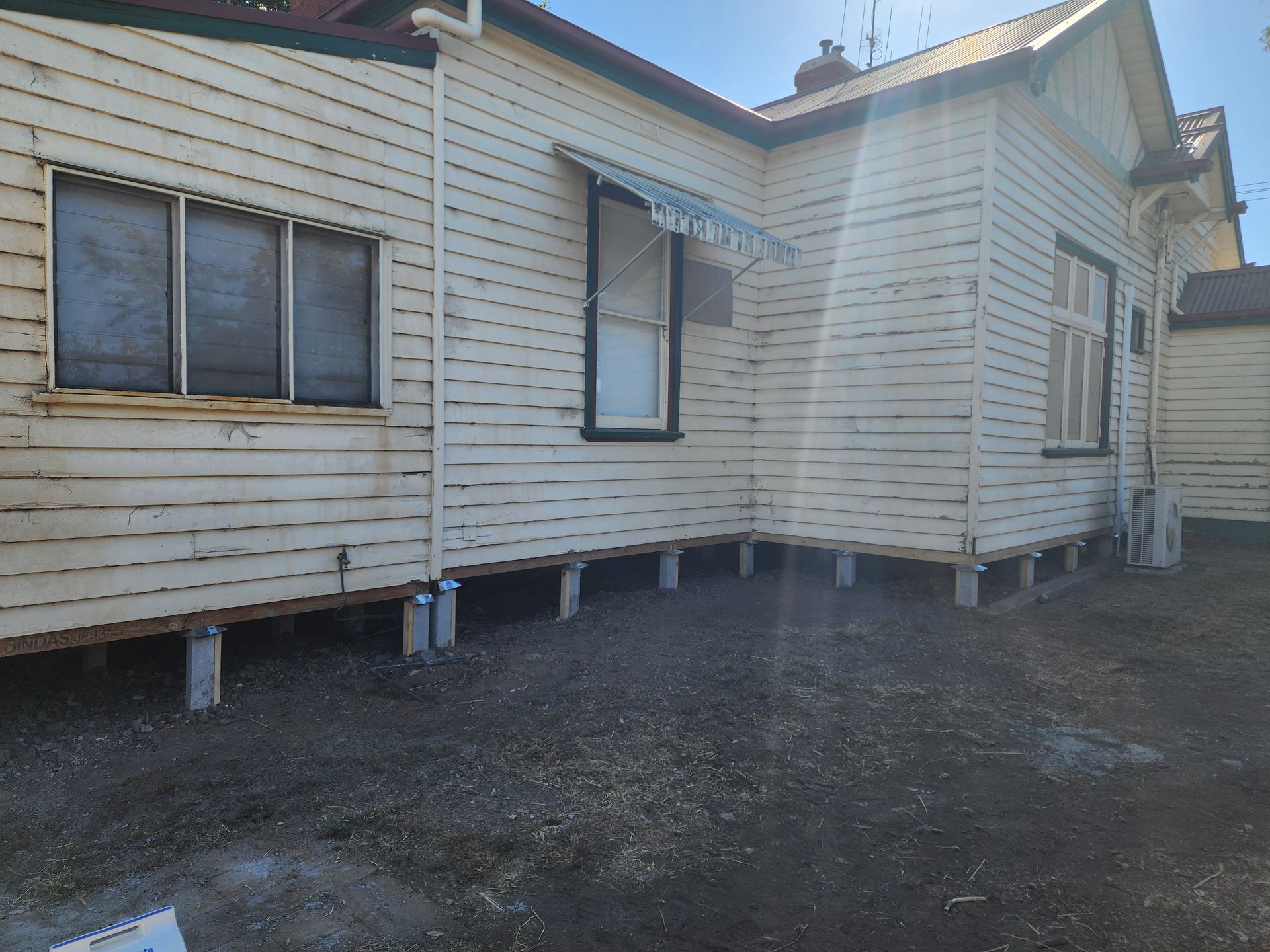 Weathered white house exterior on stilts with windows and a dirt yard under clear daylight