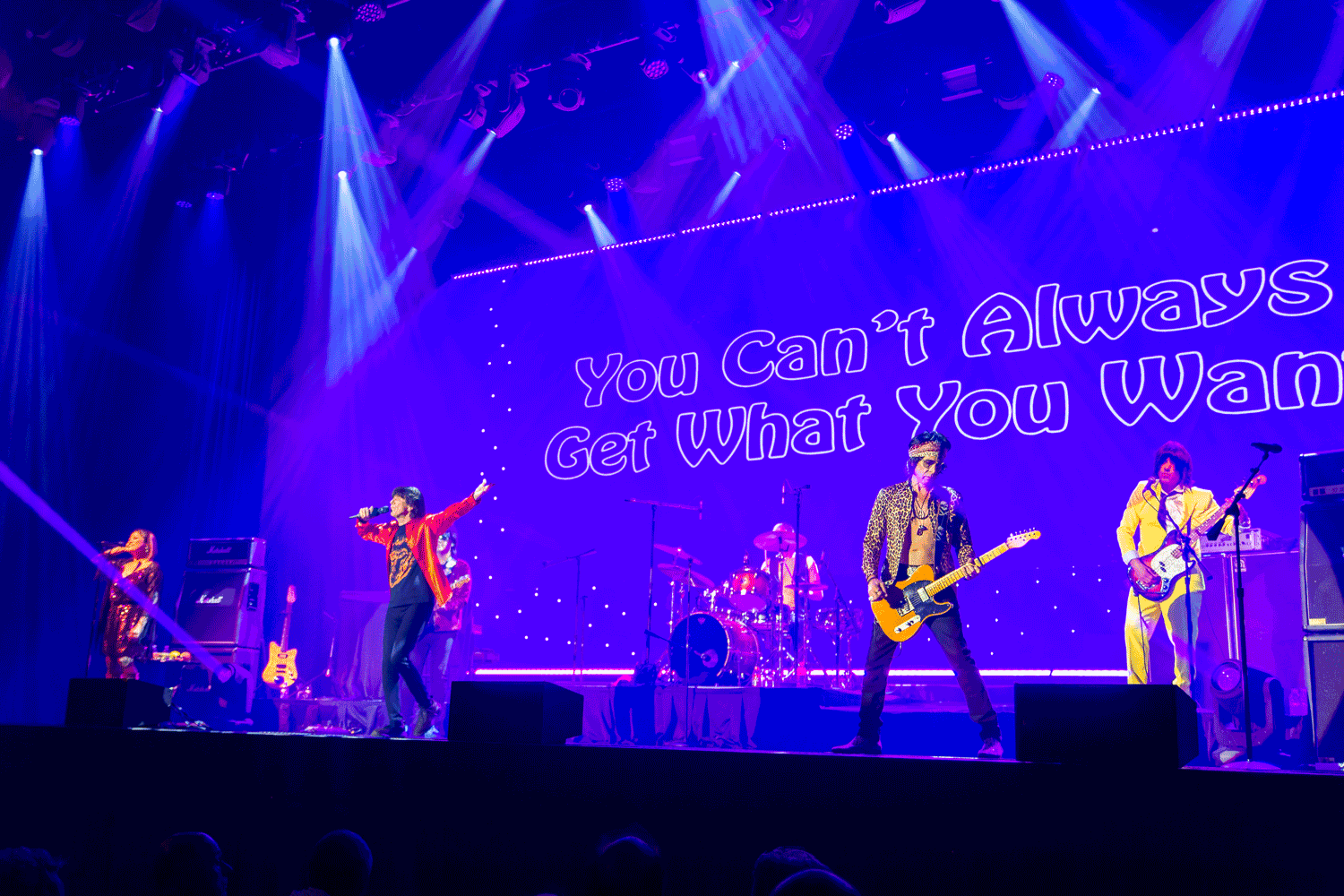 Band performing on a purple-lit stage with “You Can’t Always Get What You Want” projected behind them