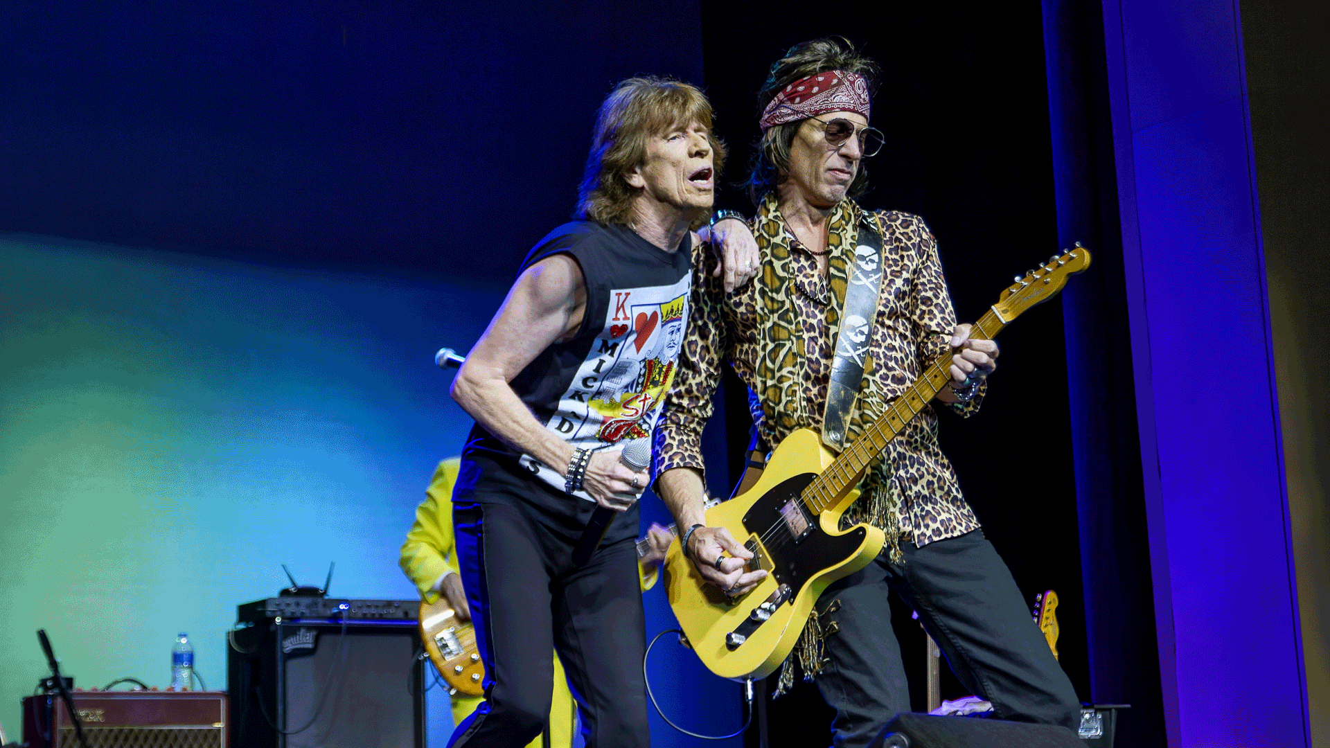 Two musicians performing on stage, one singing into a mic and one playing yellow electric guitar.