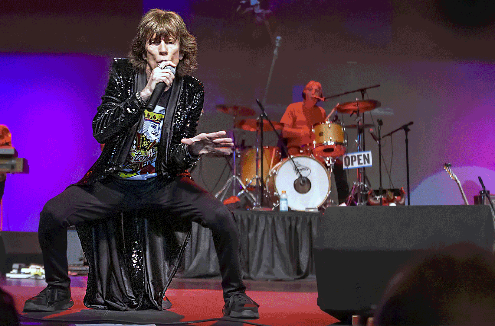 Singer in black crouches on stage under purple lights, holding a microphone while a drummer plays behind.
