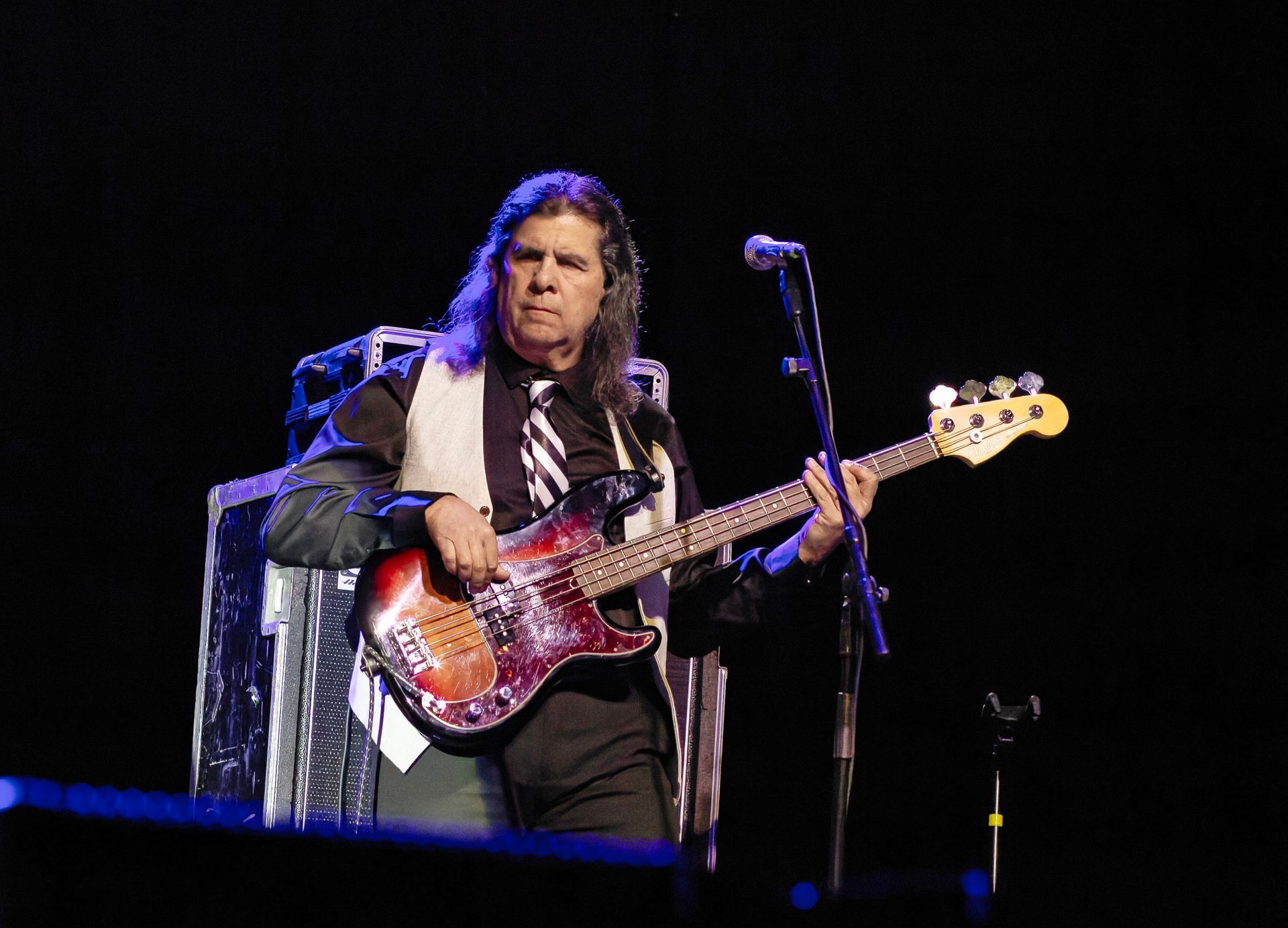 Musician playing a red electric bass onstage under blue lighting, beside a microphone stand