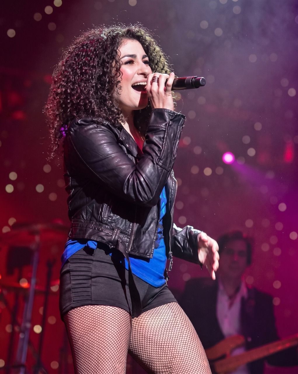 Singer performing on stage with a microphone, wearing a black jacket and fishnet tights under pink lights.