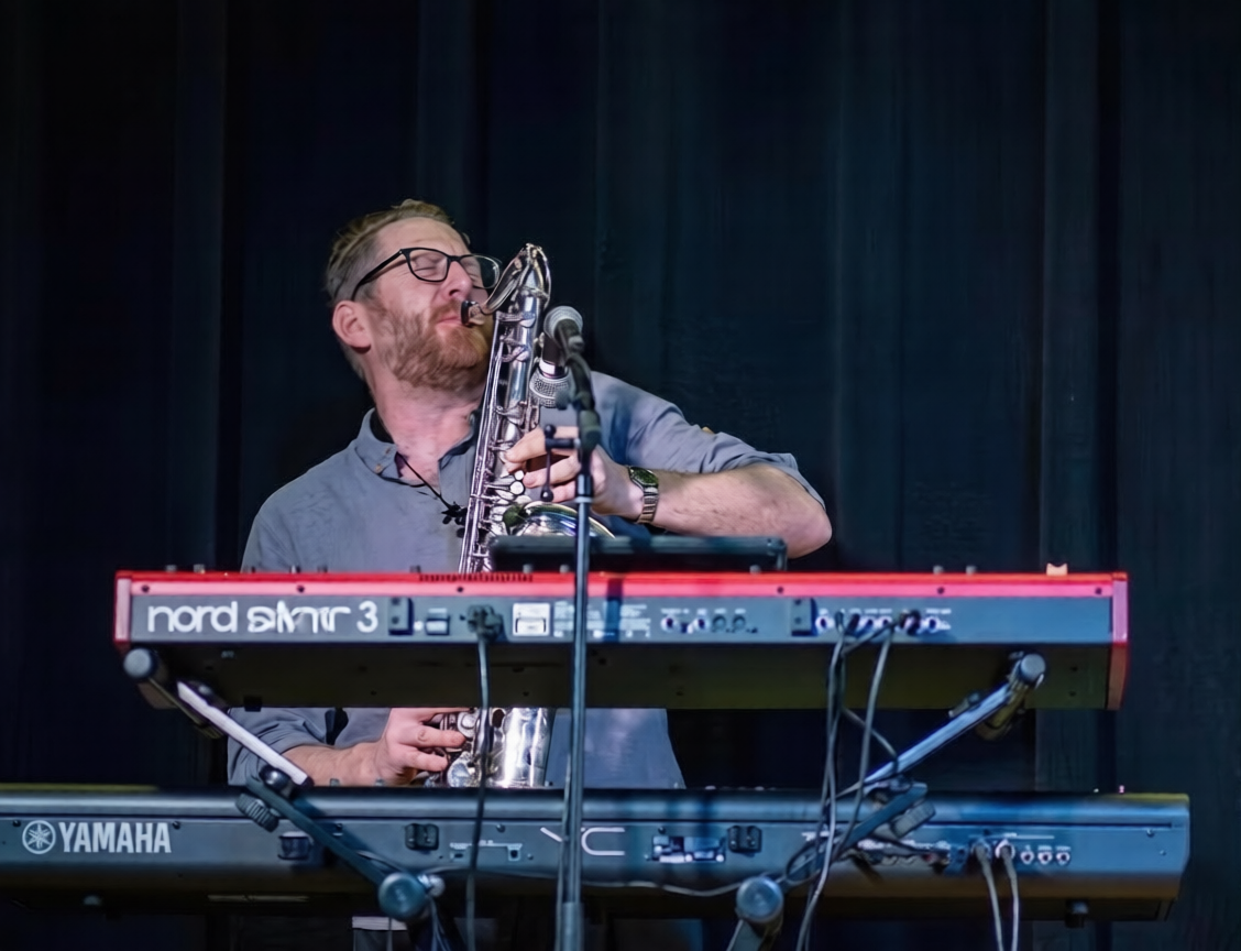 Musician playing saxophone behind red Nord keyboard on stage with dark curtains