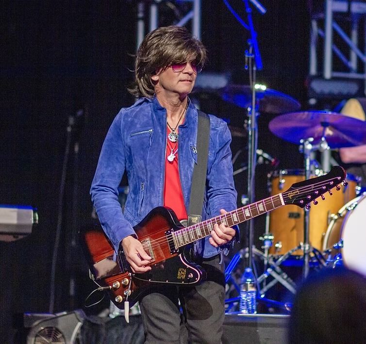 Musician playing electric guitar onstage under blue lights, wearing a blue jacket and red shirt.