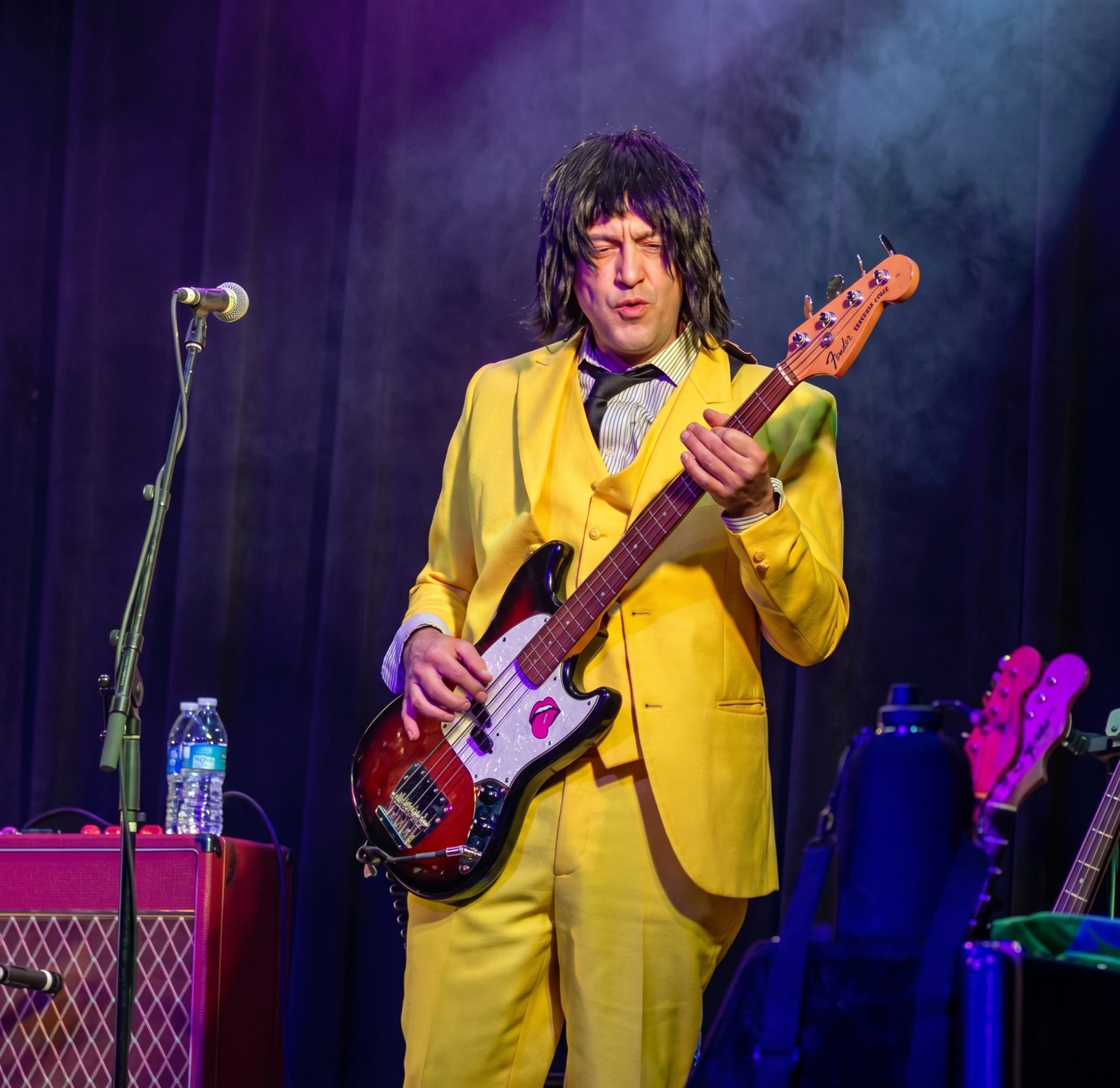 Musician in a yellow suit playing bass guitar on a lit stage with purple curtains and a microphone stand