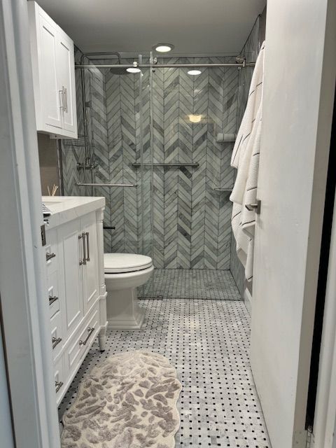 Small bathroom with gray herringbone shower tile, white cabinets, and patterned floor mat.