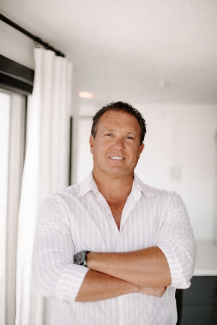 Man standing indoors with arms crossed, wearing a white shirt in a bright hallway