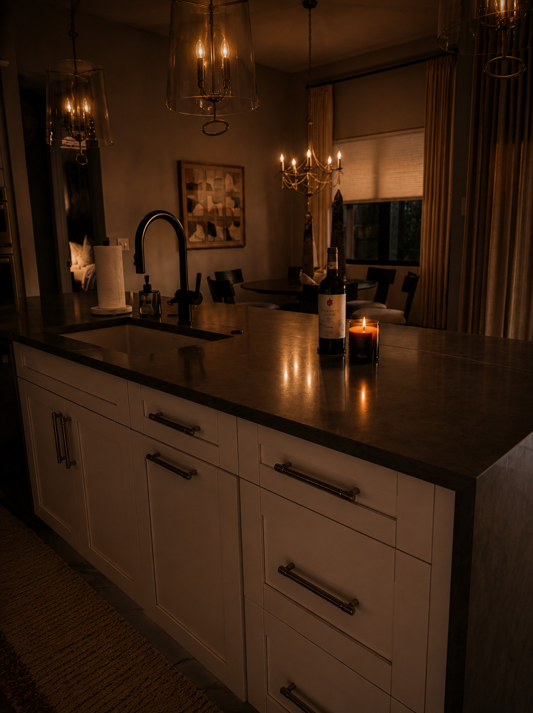 Dimly lit kitchen with white cabinets, a dark island countertop, sink, candles, and pendant lights.