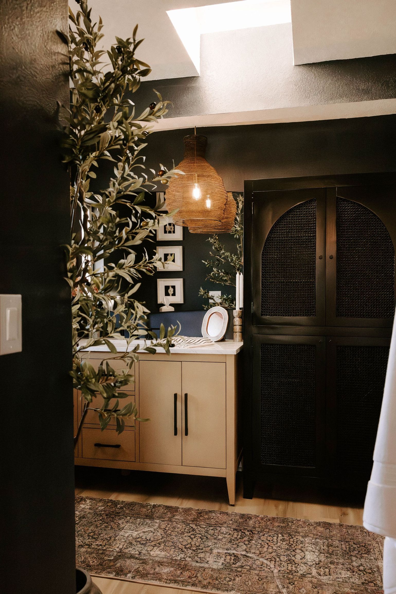 Small bathroom vanity with plant, lit mirror, and black arched cabinet doors under skylight.