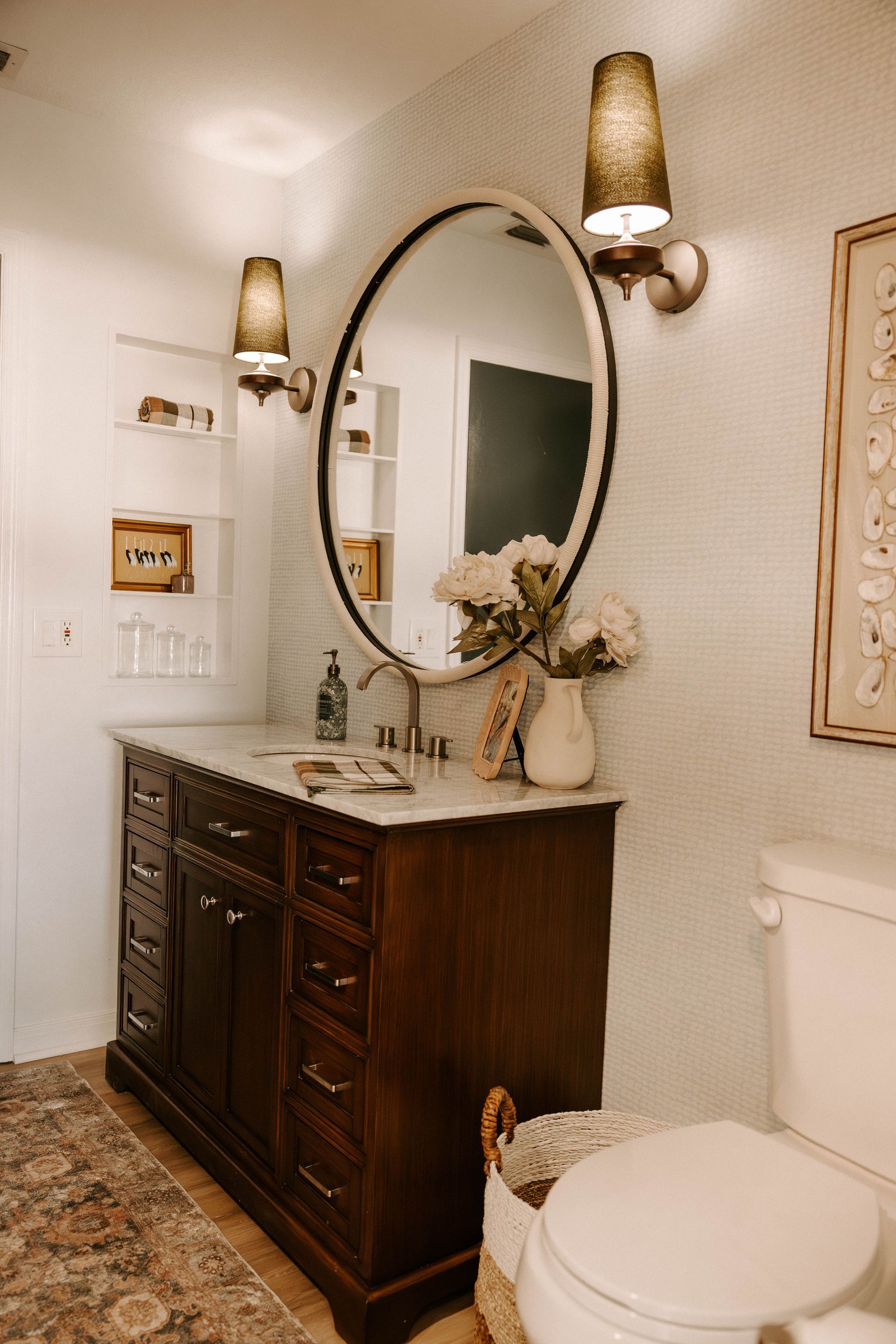 Bathroom vanity with oval mirror, wall sconces, dark wood cabinet, and white toilet against white walls.