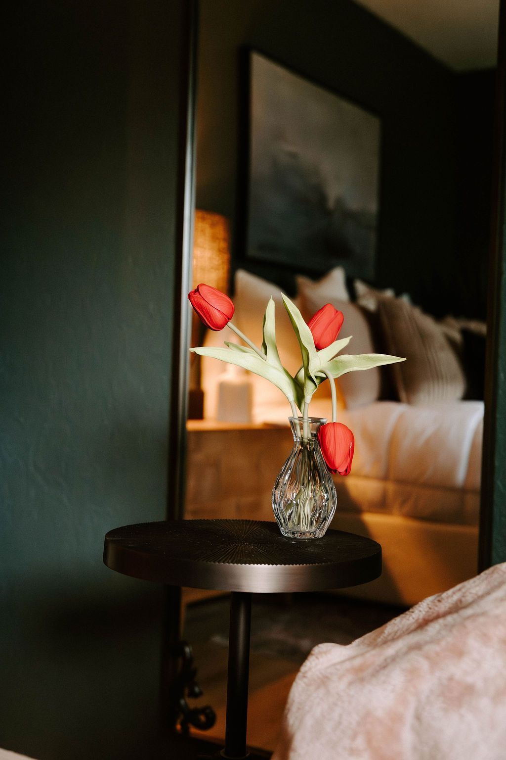 Red flowers in a small vase on a round side table beside a bed in a dim bedroom.