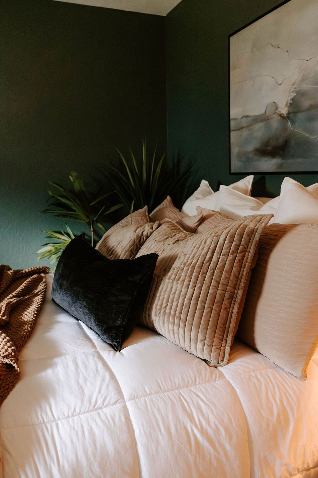 Cozy bed with white bedding and black pillows in a dark green bedroom, with a plant and abstract wall art