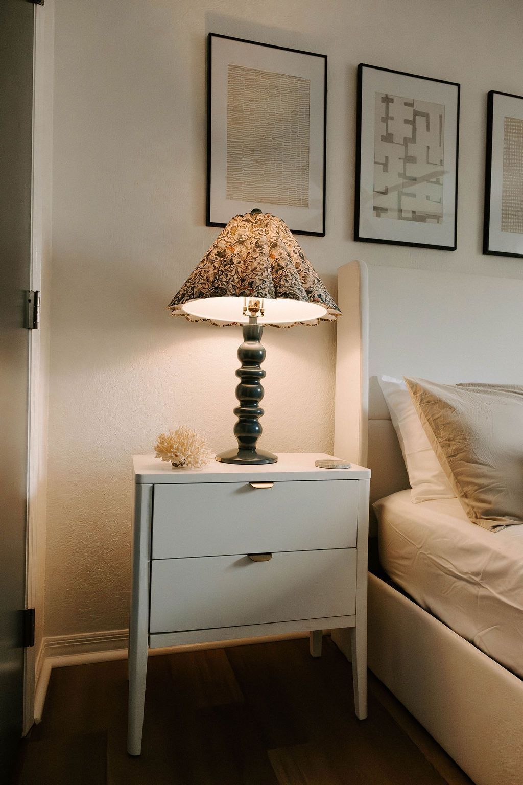 Lamp on a white bedside table beside a beige bed in a softly lit bedroom
