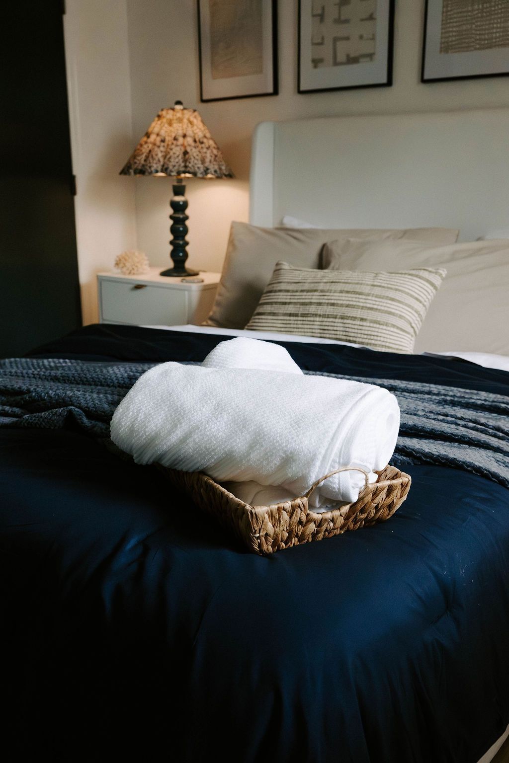 Neatly made bed with dark navy bedding, white pillows, and a towel basket at the foot in a cozy bedroom