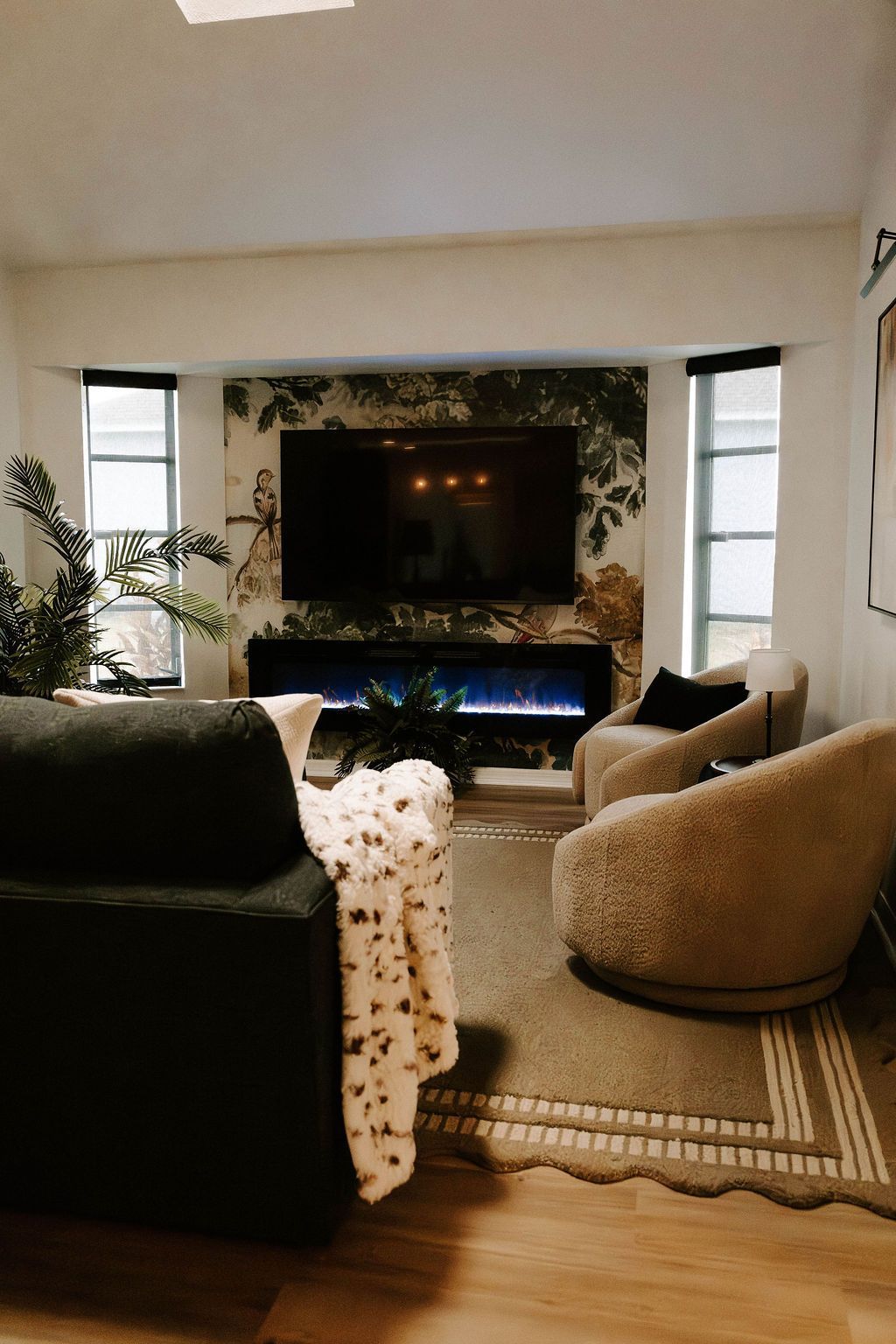Cozy living room with a TV, fireplace, plants, and a beige swivel chair on a rug.