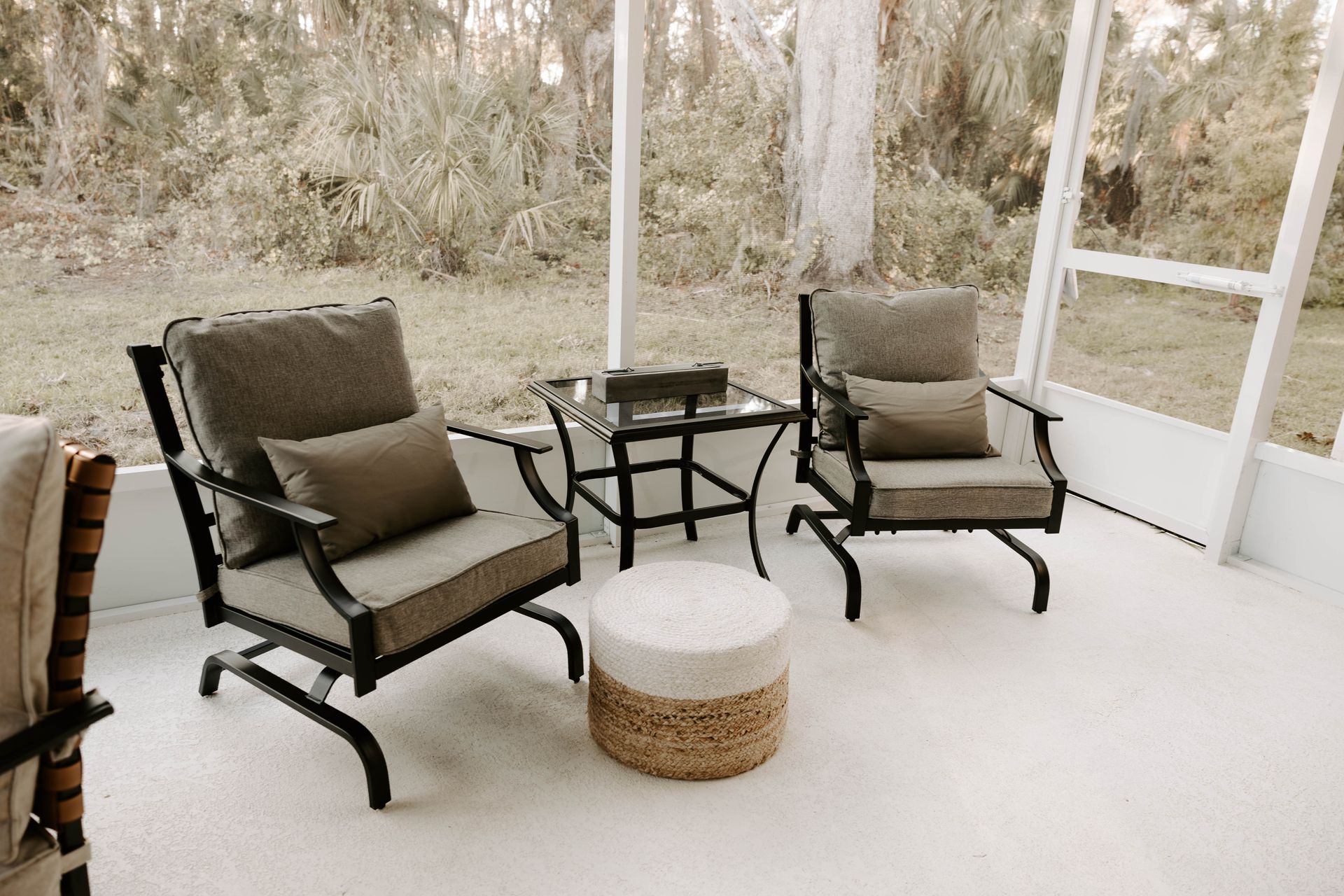 Two black lounge chairs and a wicker stool on a screened porch with a small table and wooded view.