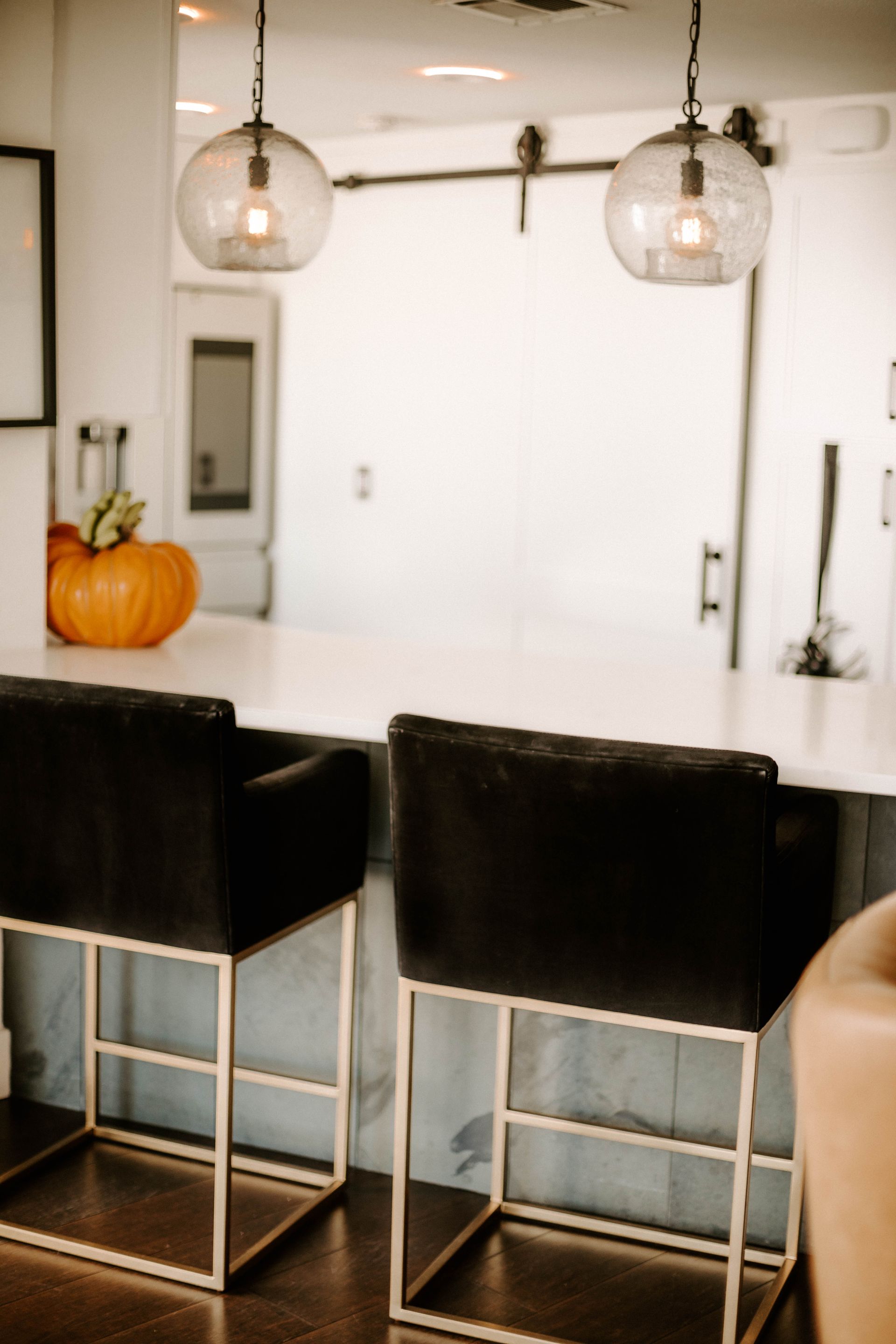 Modern kitchen with black bar stools, hanging glass pendant lights, and a pumpkin on the counter