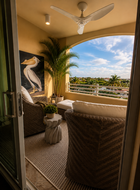 Covered balcony with wicker chairs, potted palm, ceiling fan, and ocean-view railing at sunset