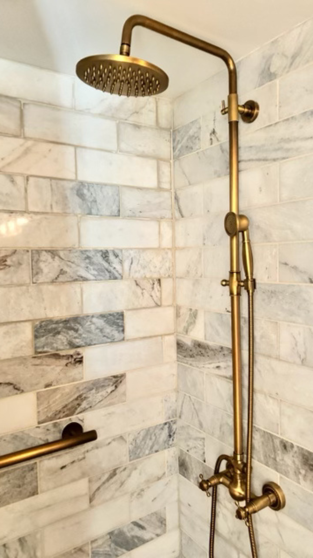 Brass rainfall shower head and handheld sprayer against a white marble tile shower wall.