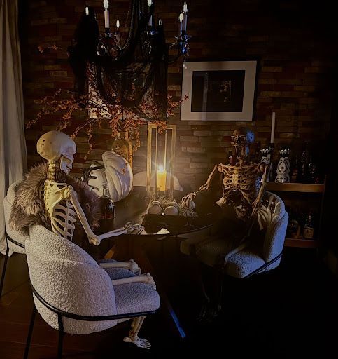 Two skeletons seated at a candlelit table in a dim, brick-walled room with a chandelier