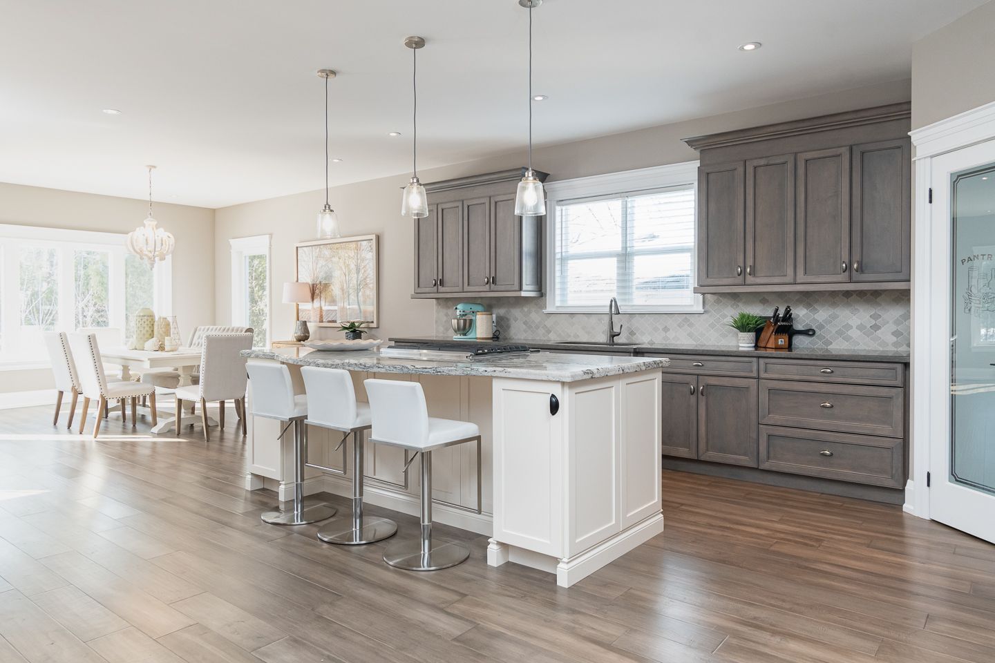 Bright modern kitchen with gray cabinets, white island stools, and an open dining area with wood floors