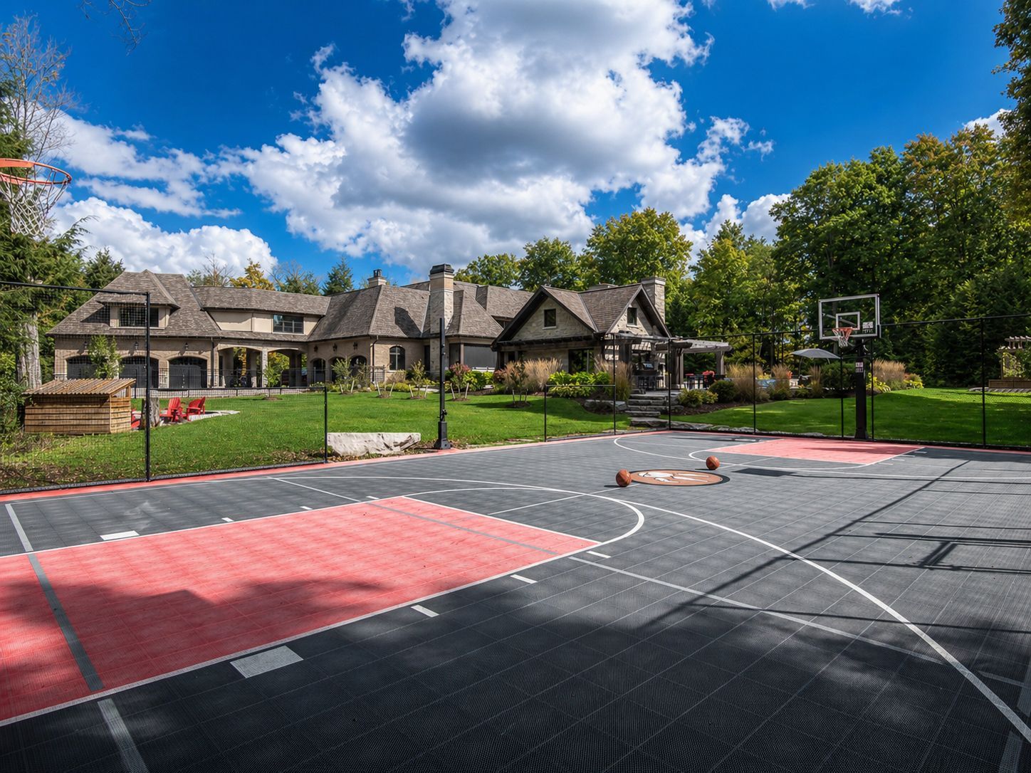 Outdoor basketball court with a house and trees in the background under a bright blue sky.
