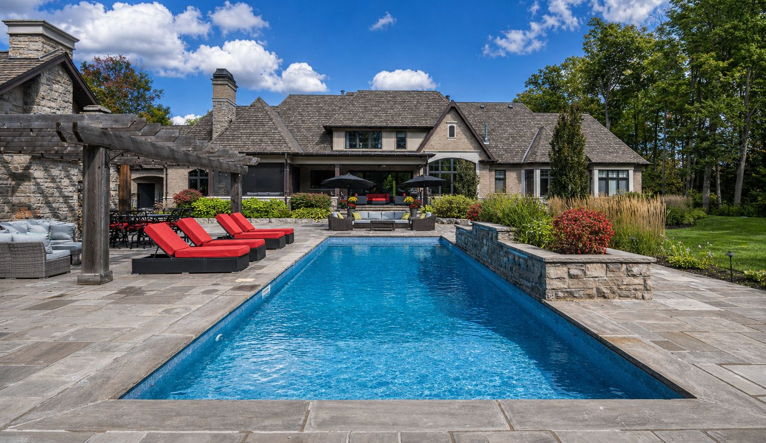 Stone patio with blue swimming pool, red lounge chairs, and a rustic house under a partly cloudy sky