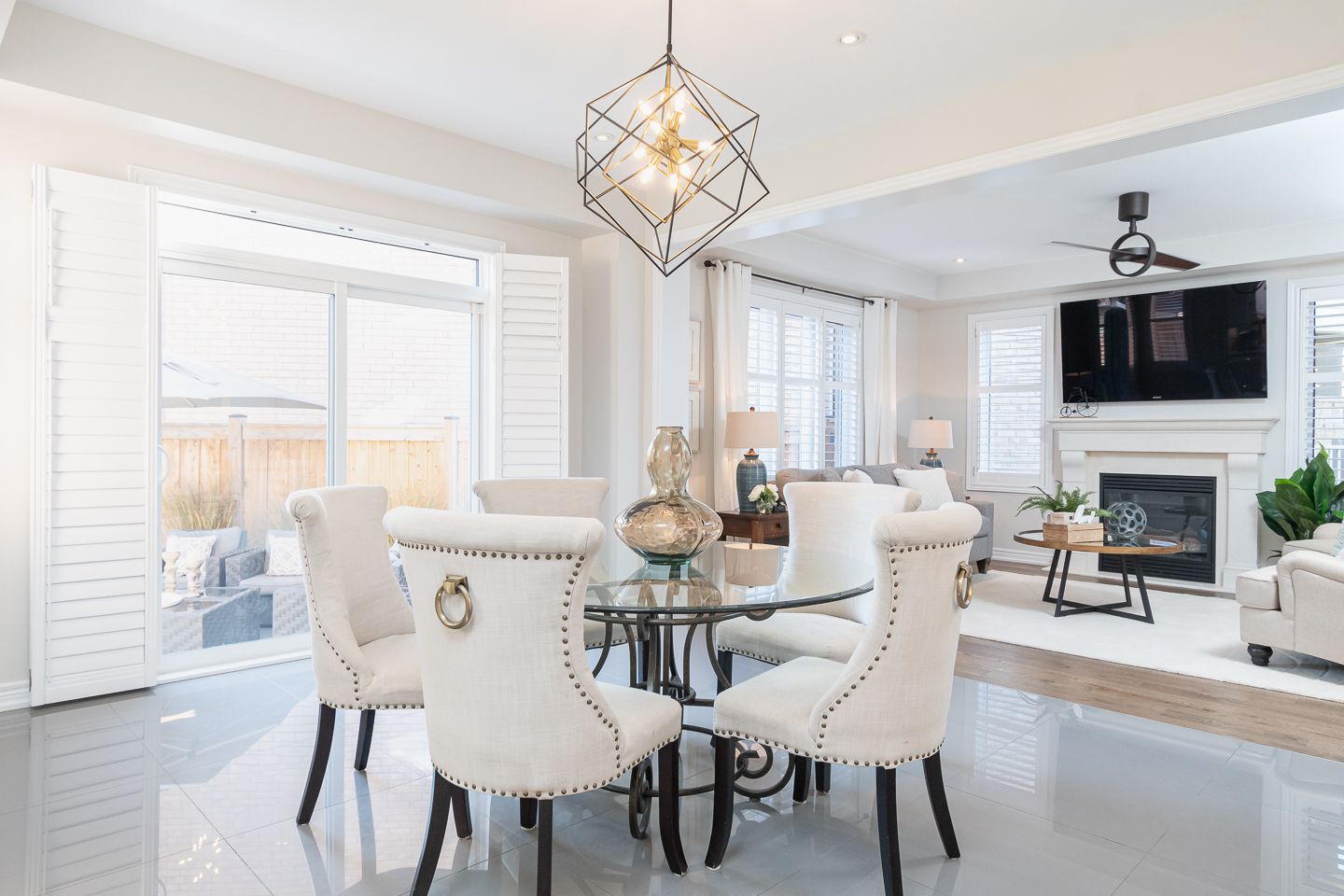 Bright open dining room with white chairs, glass table, chandelier, and TV above a fireplace