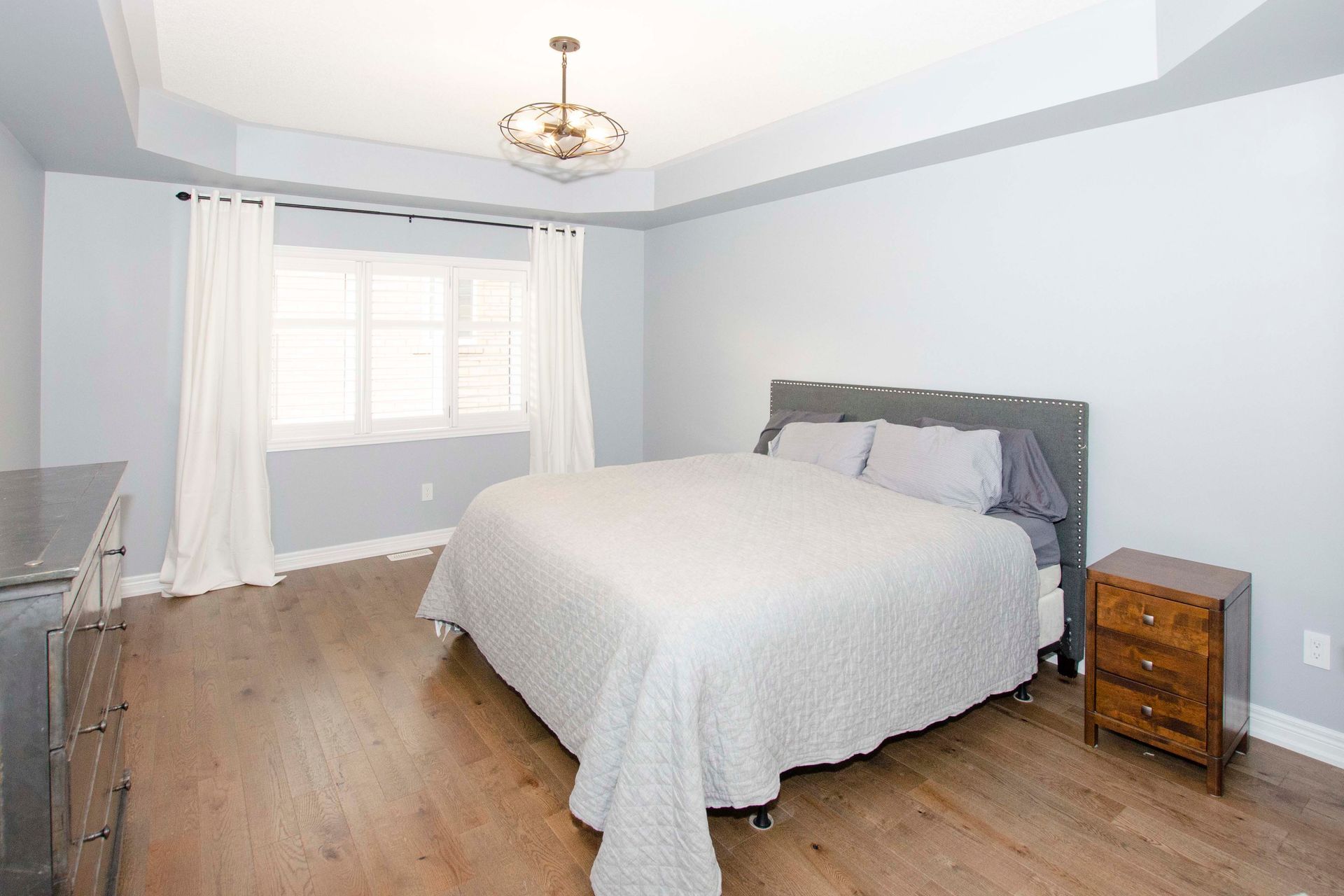 Bright bedroom with a large bed, wooden floor, window with curtains, and a small nightstand.