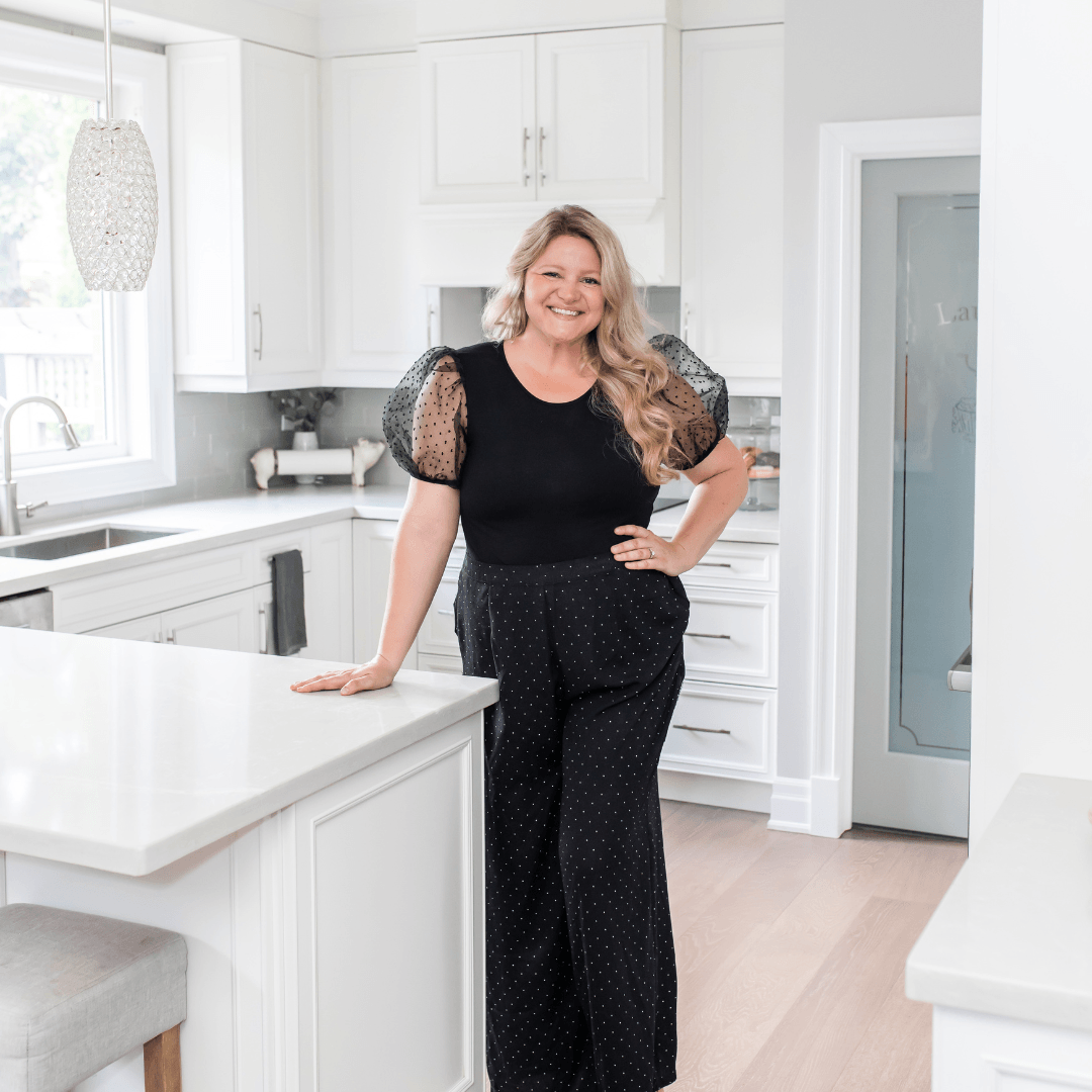 Woman leaning on a kitchen island, smiling. She's in a black top and pants, in a bright, modern kitchen.