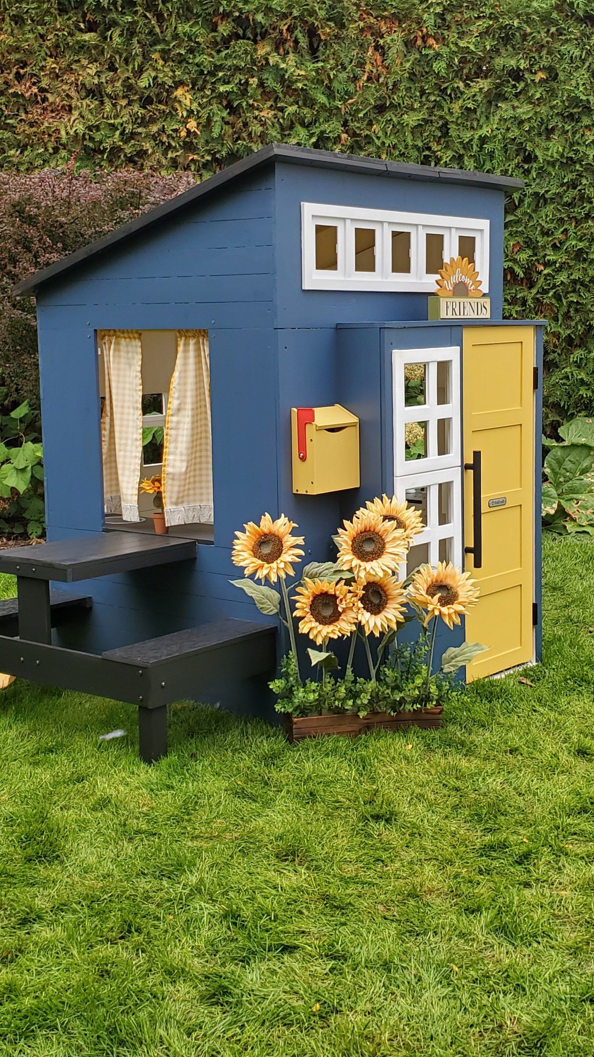 A blue and yellow playhouse with a picnic table and sunflowers in front of it.