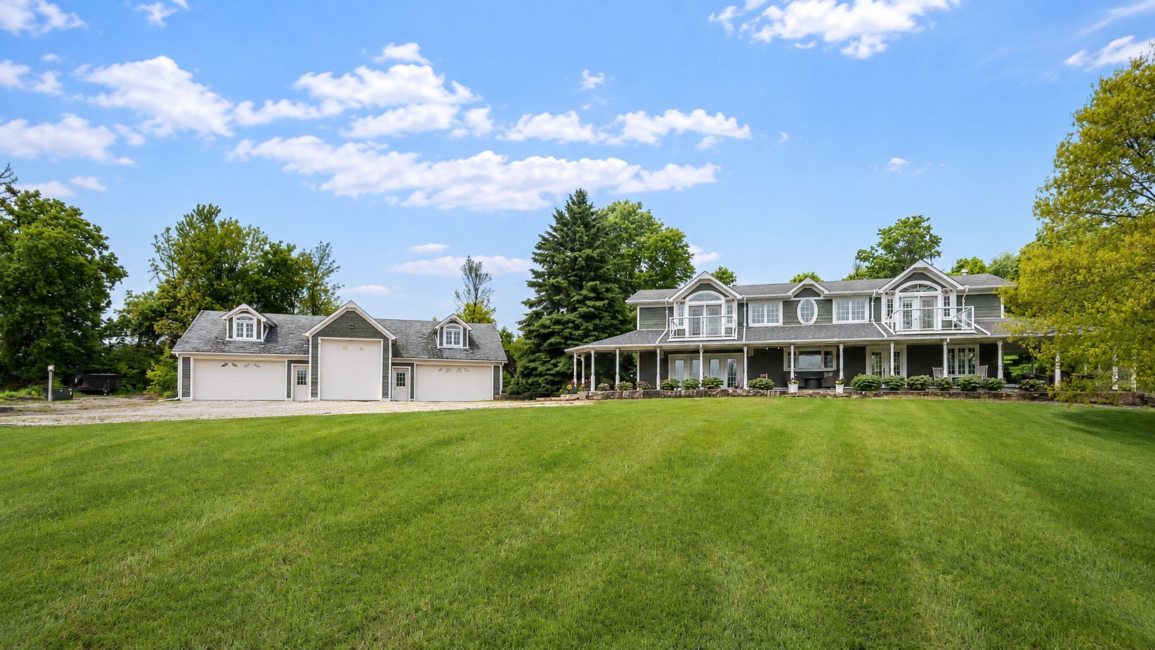 Large white farmhouse with attached garage on a wide green lawn under a blue sky with clouds