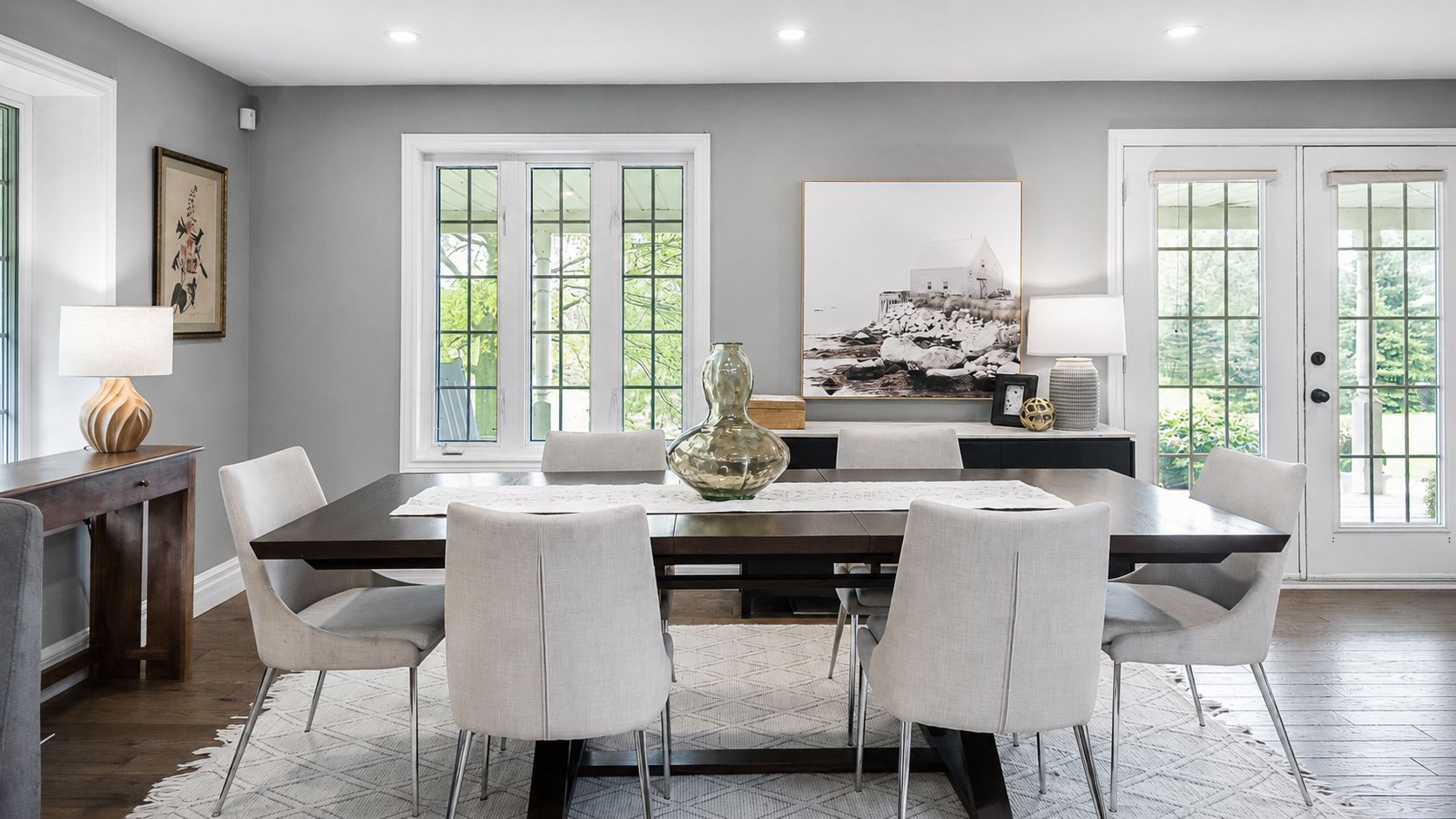 Modern dining room with a long table, white chairs, gray walls, and large windows with garden views