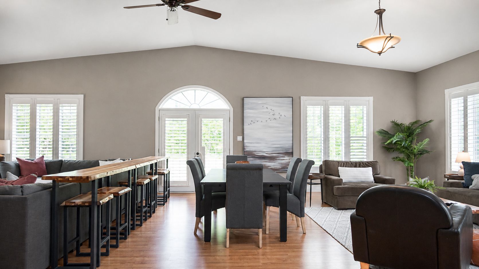 Bright open dining and living room with gray walls, wood floors, black chairs, and a large arched window.