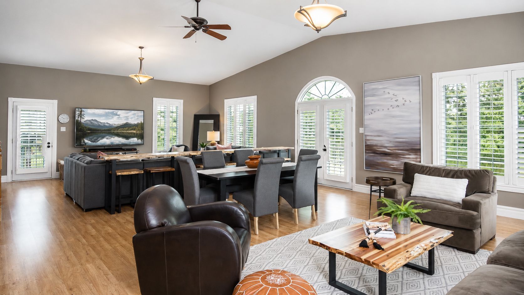 Bright open living room with gray sofa, black recliners, TV, and dining area beside large windows