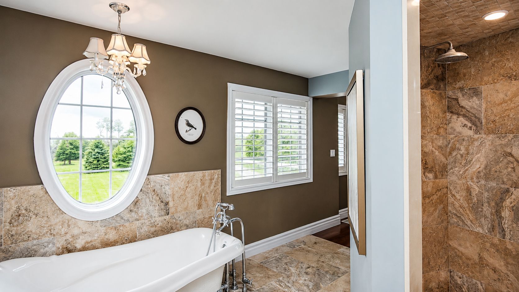 Elegant bathroom with oval window, freestanding tub, chandelier, and beige stone tile shower
