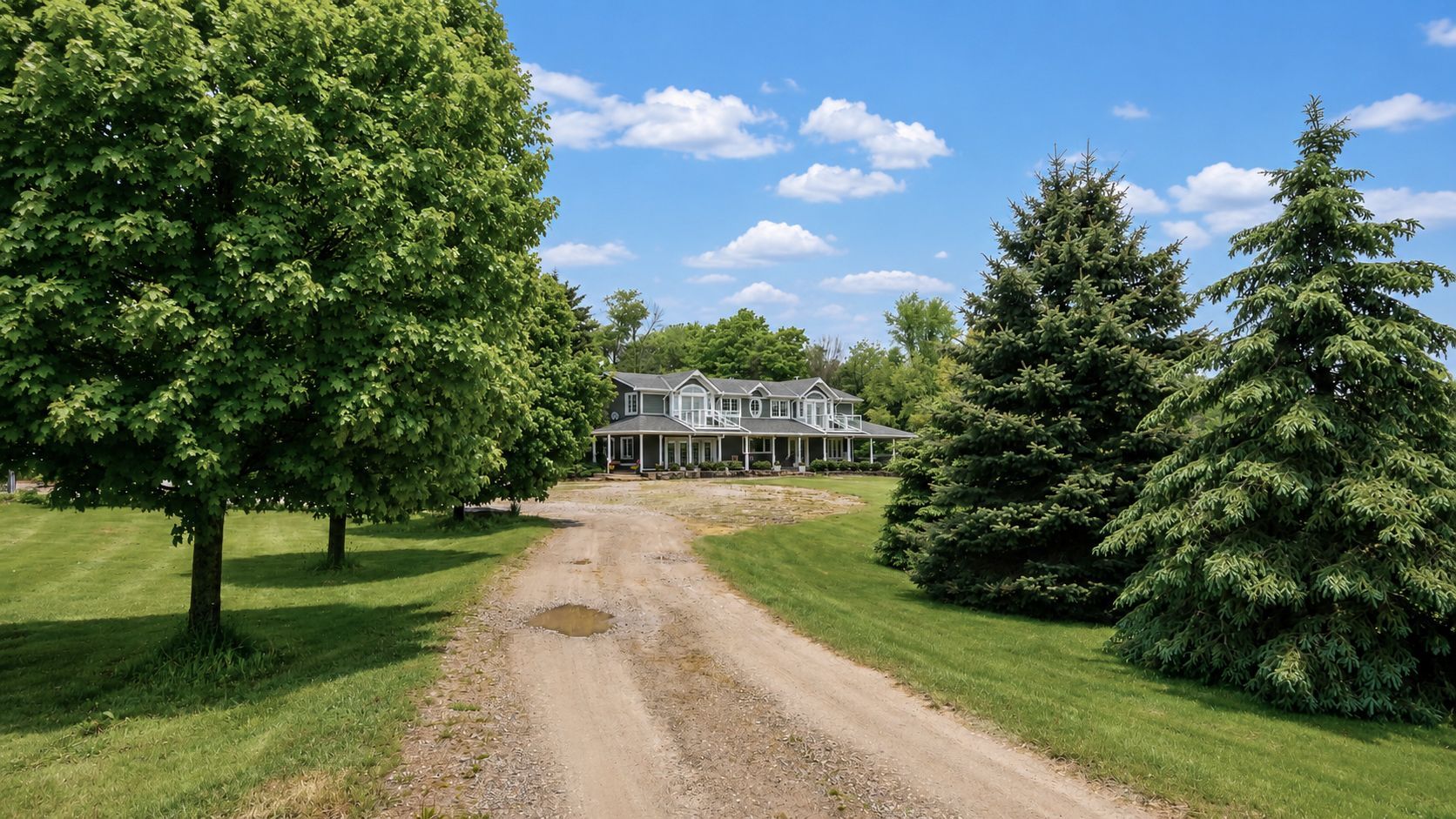 Gravel driveway leading to a white house, framed by green trees under a bright blue sky.