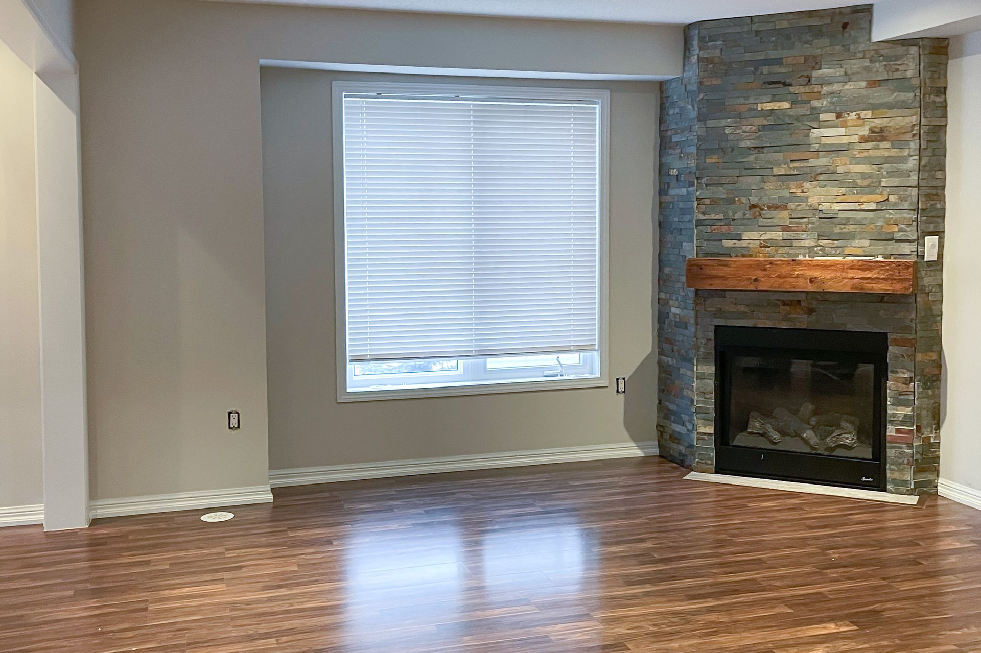 Empty living room with wood floors, gray walls, a window, and a stone fireplace.