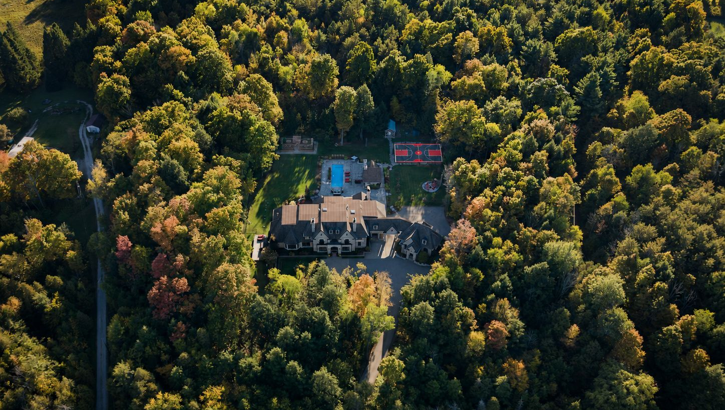 Aerial view of a large house with a pool, surrounded by dense green forest and a few nearby buildings.