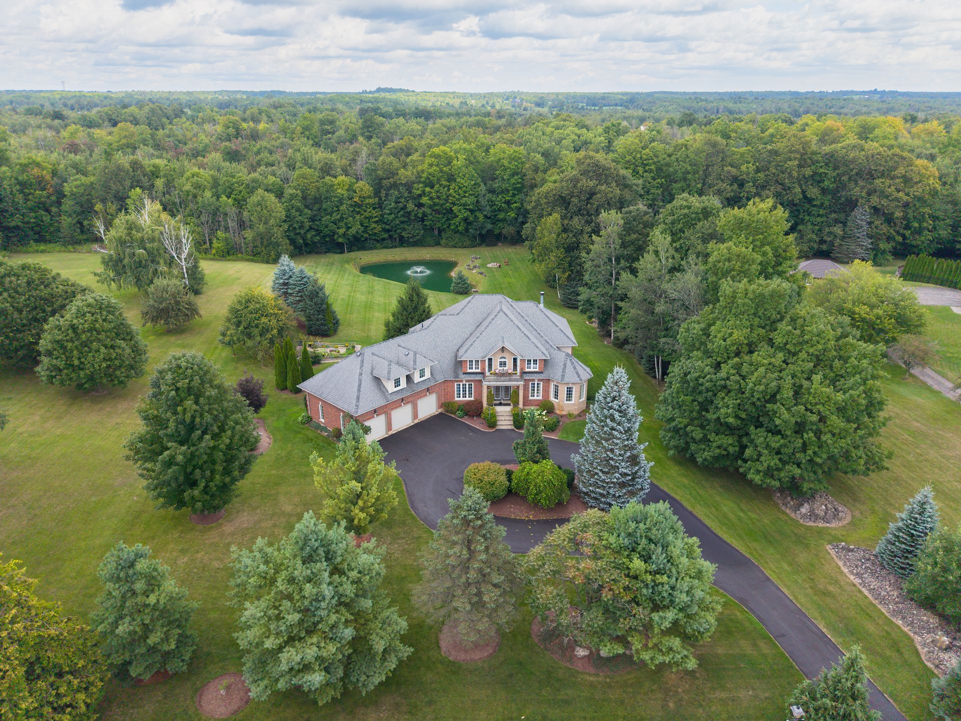 Aerial view of a large house surrounded by trees, lawns, and a driveway in a wooded rural area