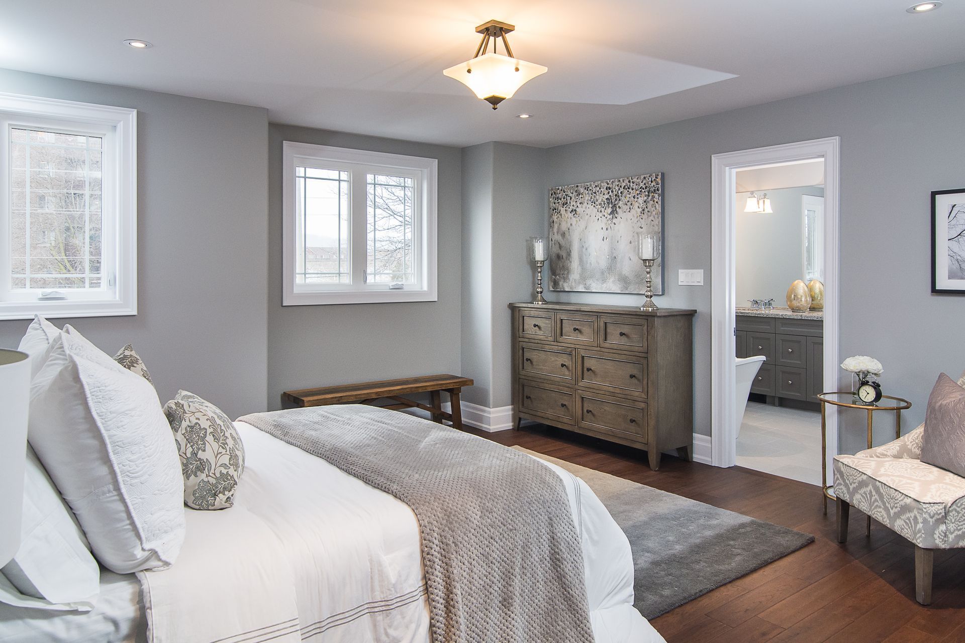 Staged bedroom with gray walls, wooden floor, bed, dresser, and access to a bathroom.