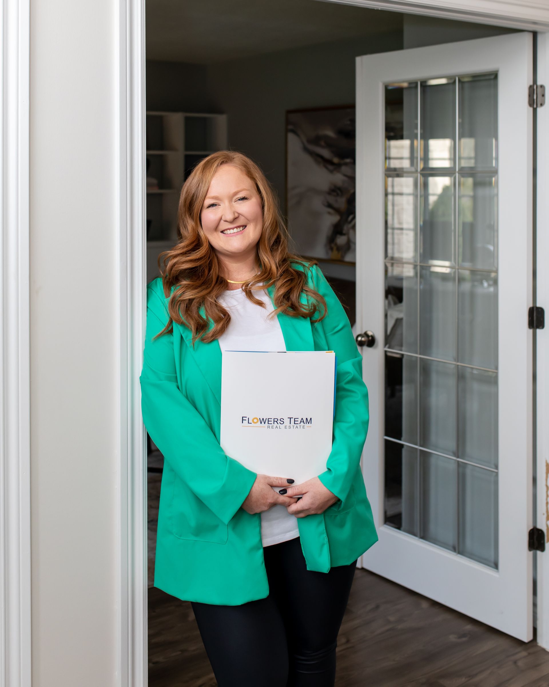 Woman in green blazer smiles, holding paperwork in doorway.