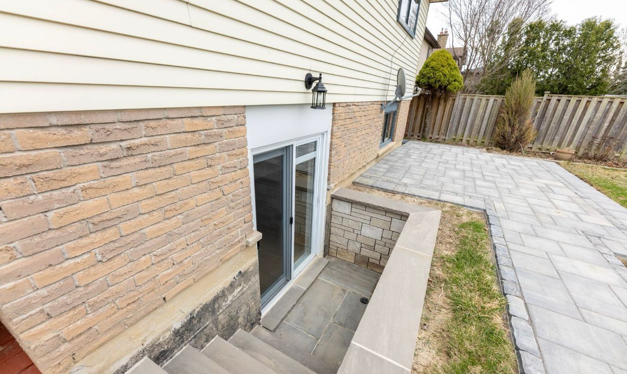 Basement stairs beside house with brick wall, concrete retaining wall, and paved patio leading to a backyard fence