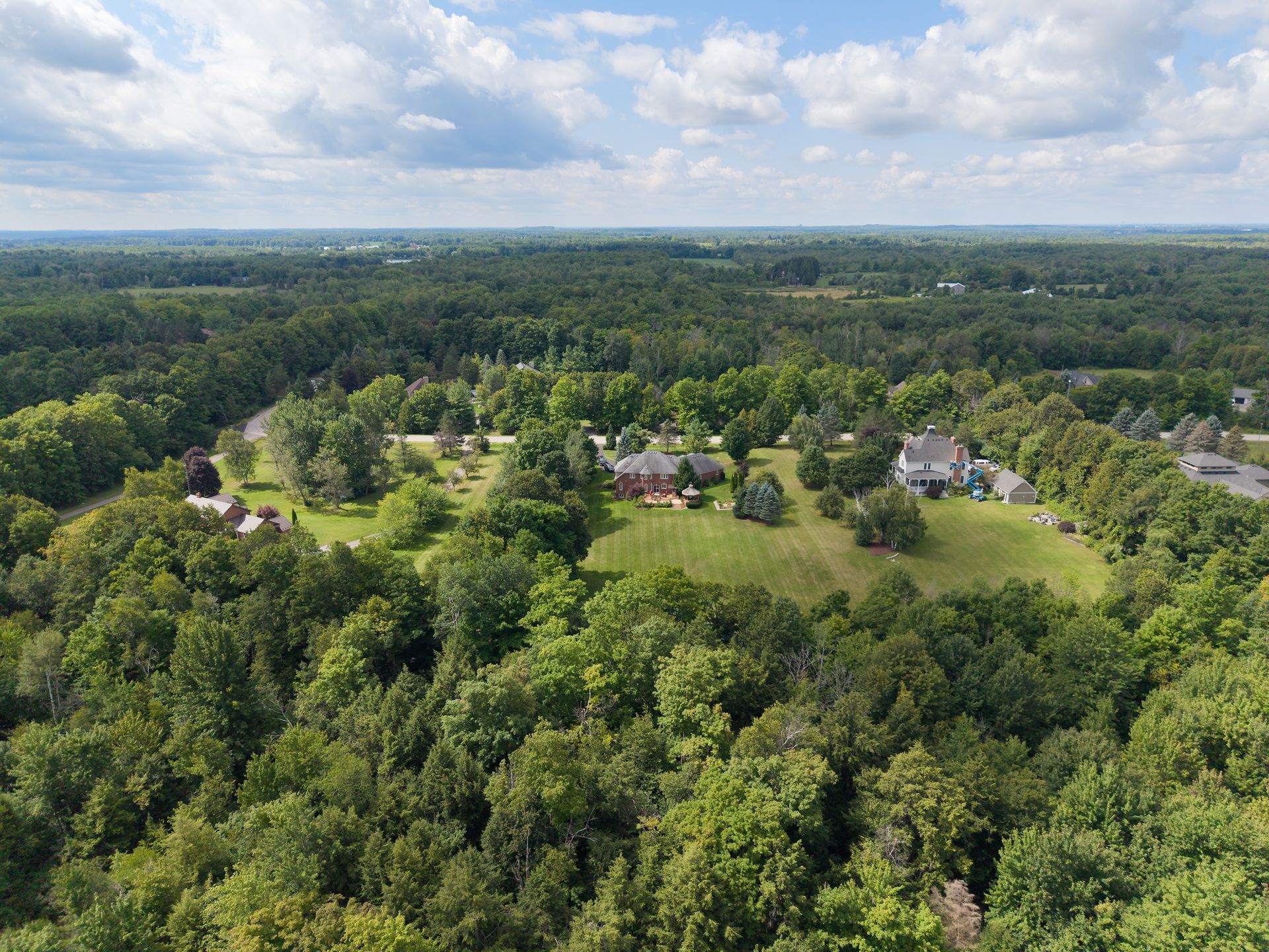 Aerial view of a forested neighborhood with homes and lawns amid dense green trees under a cloudy sky