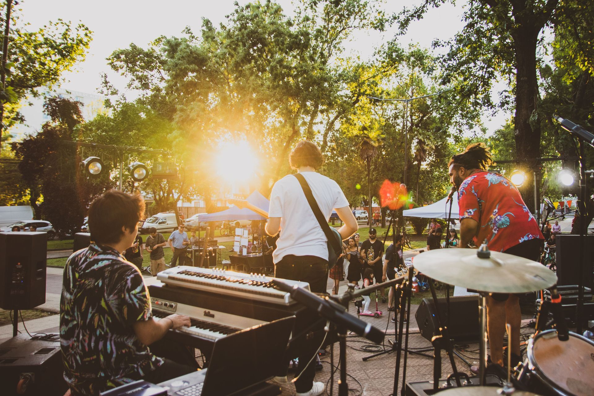 Live outdoor band performing at sunset with keyboards, drums, guitars, and bright sun through trees